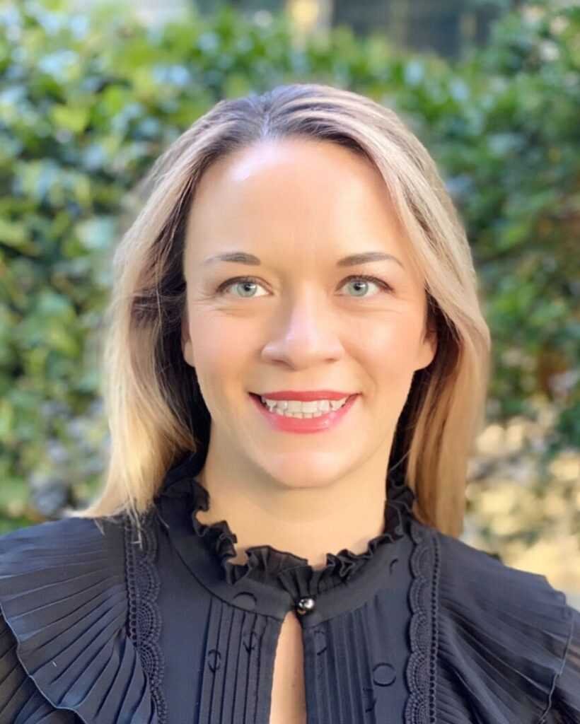 A smiling, blue-eyed Tessa Bailey with light hair poses outdoors in front of lush greenery, her black ruffle blouse echoing the playful charm of "Pitcher Perfect by Tessa Bailey (Big Shots, 4 of 5).