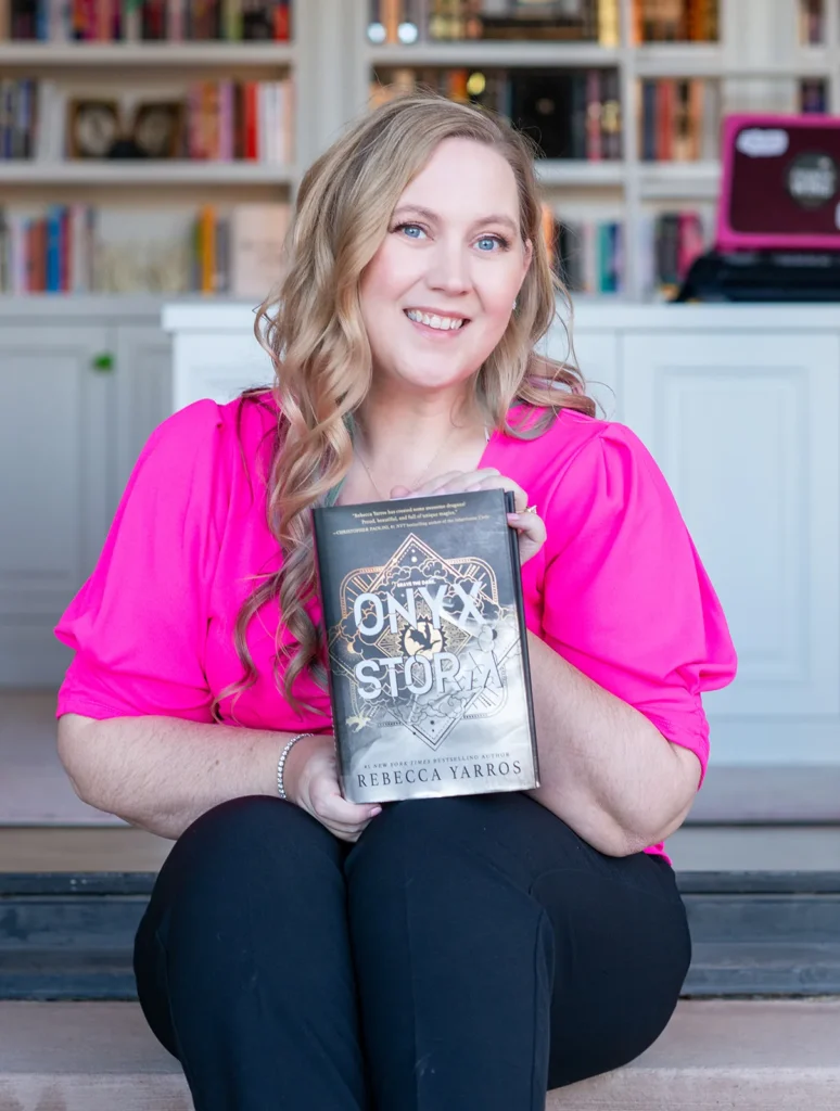 Rebecca Yarros sits indoors holding The Last Letter by Rebecca Yarros, with bookshelves visible in the background.