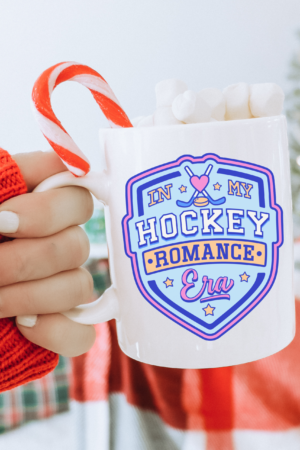 A hand in a red-and-white striped sleeve holds the Hockey Romance Era Bookish Coffee Mug with “In My Hockey Romance Era” on it, filled with marshmallows and a candy cane. A glowing Christmas tree adds festive charm—ideal for contemporary romance novel fans.