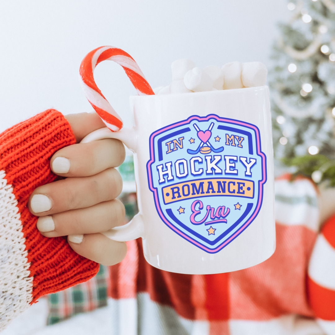 A hand in a red-and-white striped sleeve holds the Hockey Romance Era Bookish Coffee Mug with “In My Hockey Romance Era” on it, filled with marshmallows and a candy cane. A glowing Christmas tree adds festive charm—ideal for contemporary romance novel fans.