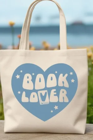 The Book Lover Tote is a cream tote bag with a large blue heart, playful “BOOK LOVER” text, stars, an open book, flowers, and a seascape—perfect for fans of trending BookTok books.