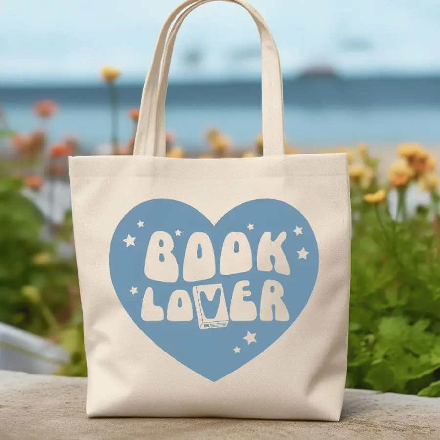The Book Lover Tote is a cream tote bag with a large blue heart, playful “BOOK LOVER” text, stars, an open book, flowers, and a seascape—perfect for fans of trending BookTok books.
