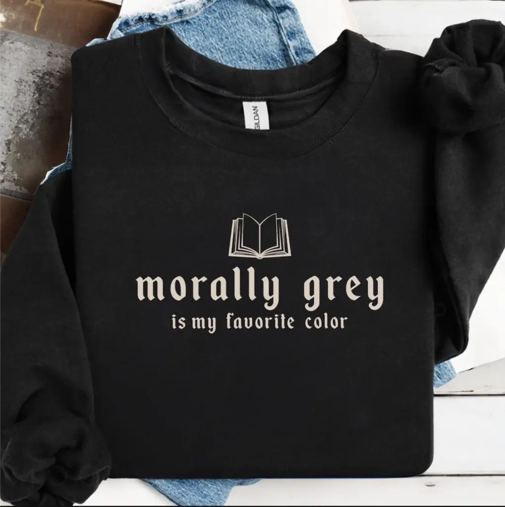 The Morally Grey Sweatshirt, folded on light blue denim jeans, features an open book illustration and beige "morally grey is my favorite color" text—a stylish nod to contemporary fiction and viral book recommendations.