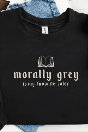 The Morally Grey Sweatshirt, folded on light blue denim jeans, features an open book illustration and beige "morally grey is my favorite color" text—a stylish nod to contemporary fiction and viral book recommendations.