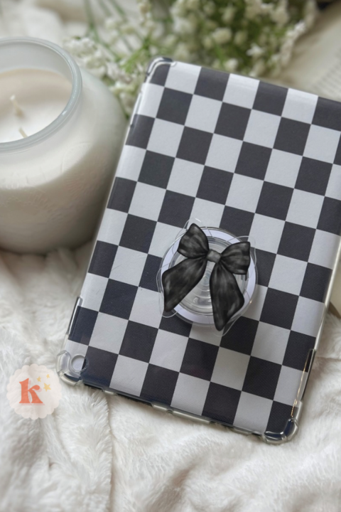 A Kindle with a Black Bow Coquette Grip rests on a white blanket, beside an open Contemporary Fiction book and a lit white candle in a glass jar.
