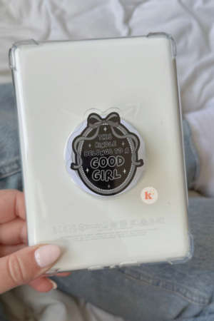 A hand with light pink nails holds a white Kindle with a MagSafe Good Girl Kindle Grip. In the background, someone in blue jeans and white socks sits on a bed, perhaps browsing Trending BookTok Books.