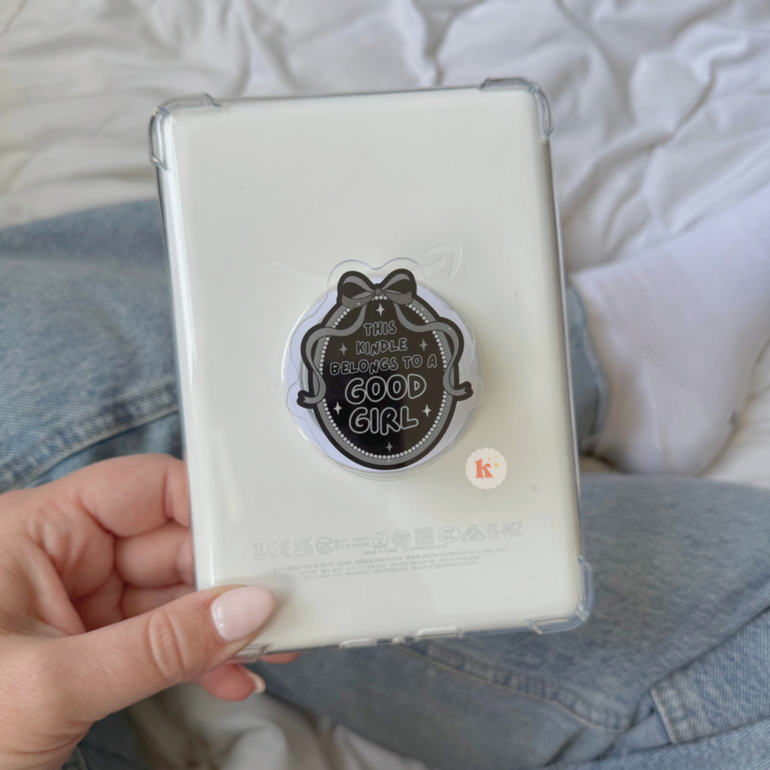 A hand with light pink nails holds a white Kindle with a MagSafe Good Girl Kindle Grip. In the background, someone in blue jeans and white socks sits on a bed, perhaps browsing Trending BookTok Books.