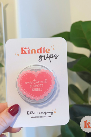 A hand holds the MagSafe Emotional Support Kindle Grip | Coquette Kindle Grip | Kobo, shaped like a pink heart and labeled “emotional SUPPORT KINDLE,” ideal for cozy reading with contemporary fiction or romance novels among green plants and soft lighting.