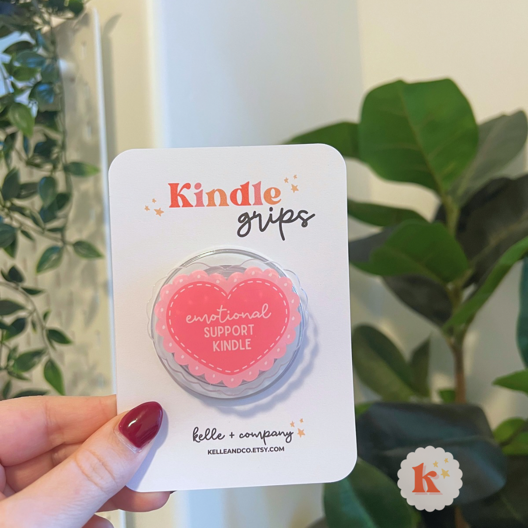 A hand holds the MagSafe Emotional Support Kindle Grip | Coquette Kindle Grip | Kobo, shaped like a pink heart and labeled “emotional SUPPORT KINDLE,” ideal for cozy reading with contemporary fiction or romance novels among green plants and soft lighting.