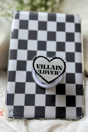 A Kindle with a black and white checkerboard case and a heart-shaped grip reading "VILLAIN + LOVER" (Kindle accessories | bookish kindle grip | kindle case stand) rests on white fabric beside an open contemporary fiction book and a lit white candle.