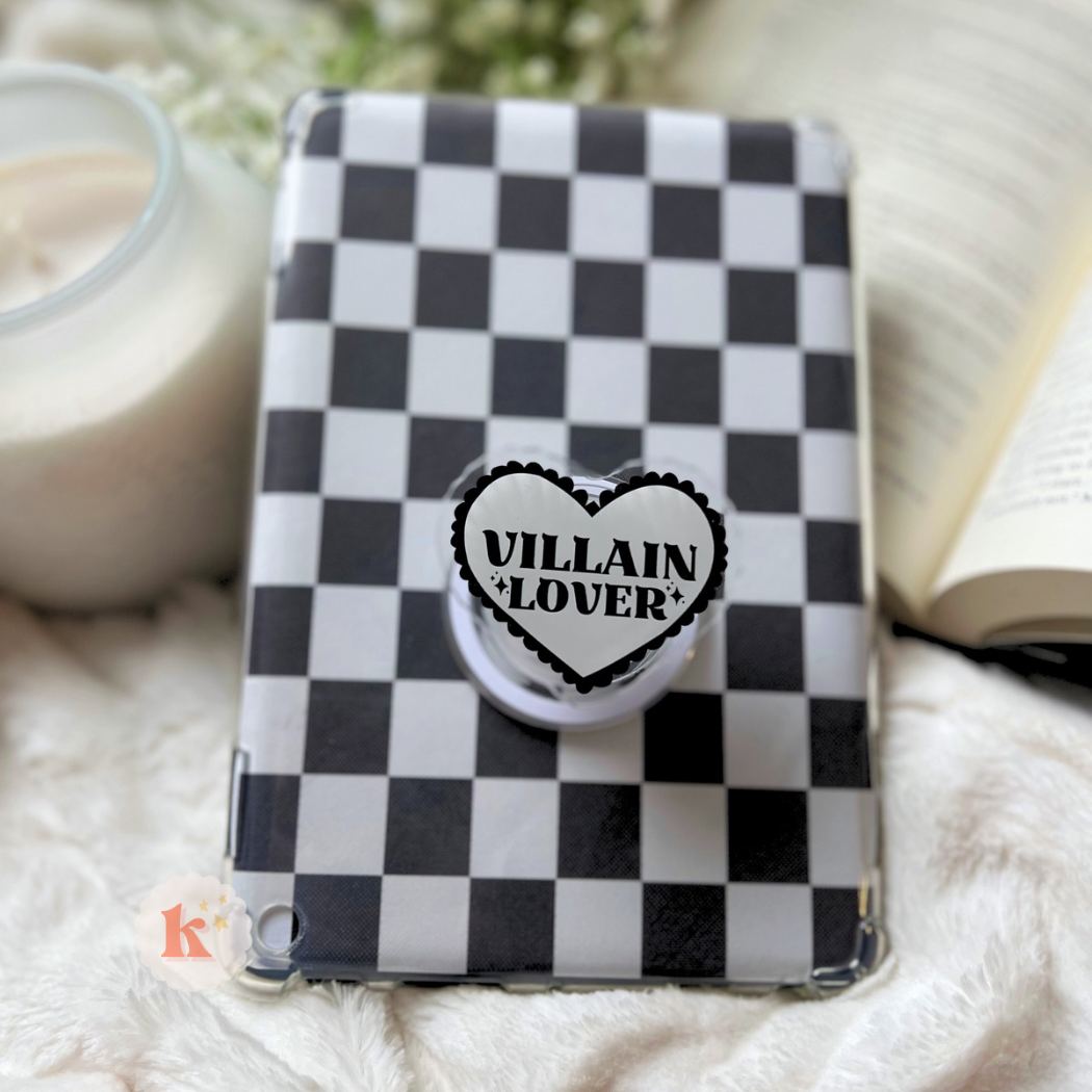 A Kindle with a black and white checkerboard case and a heart-shaped grip reading "VILLAIN + LOVER" (Kindle accessories | bookish kindle grip | kindle case stand) rests on white fabric beside an open contemporary fiction book and a lit white candle.