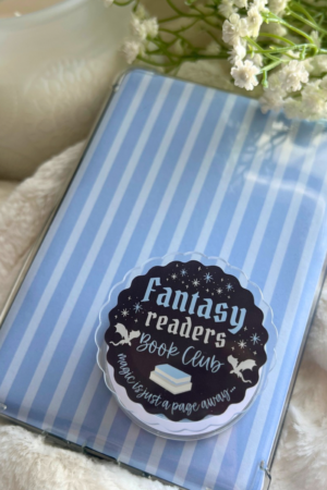 A Kindle with a blue and white striped cover and a "Fantasy readers" bookish grip rests on a fluffy surface. Flowers, a white candle, and Trending BookTok Books complete this cozy setup for fantasy lovers.
