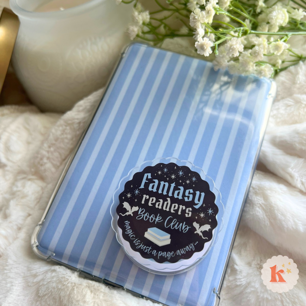 A Kindle with a blue and white striped cover and a "Fantasy readers" bookish grip rests on a fluffy surface. Flowers, a white candle, and Trending BookTok Books complete this cozy setup for fantasy lovers.