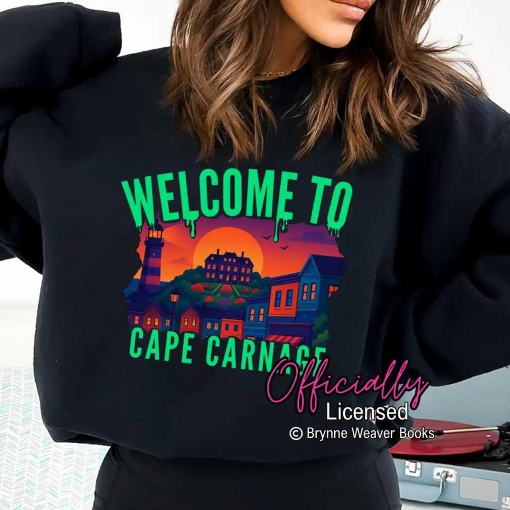 A woman wears the black Cape Carnage Sweatshirt (Tourist Season x Brynne Weaver), featuring “Welcome to Cape Carnage” in green above a sunset coastal town and lighthouse. Officially Licensed © Brynne Weaver Books appears at the bottom.