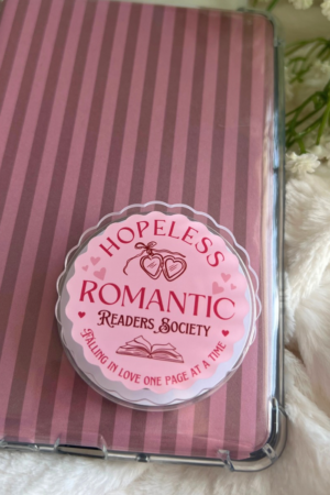 A pink "Hopeless Romantic Readers Society" badge sits on a striped notebook with flowers and a blanket—an ideal Kindle accessory and bookish grip for romance book lovers inspired by viral recommendations.