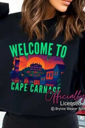 A woman wears the black Cape Carnage Sweatshirt (Tourist Season x Brynne Weaver), featuring “Welcome to Cape Carnage” in green above a sunset coastal town and lighthouse. Officially Licensed © Brynne Weaver Books appears at the bottom.