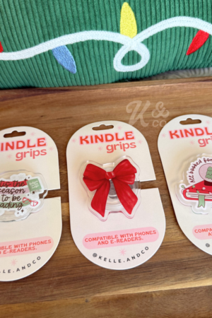 Three Christmas Kindle Grips sit on a tray, featuring a holiday book, red bow, and cat-in-a-box designs. “Christmas Kindle Grip | Christmas Book | Kindle Case”—a perfect festive gift for book lovers and trending BookTok fans.