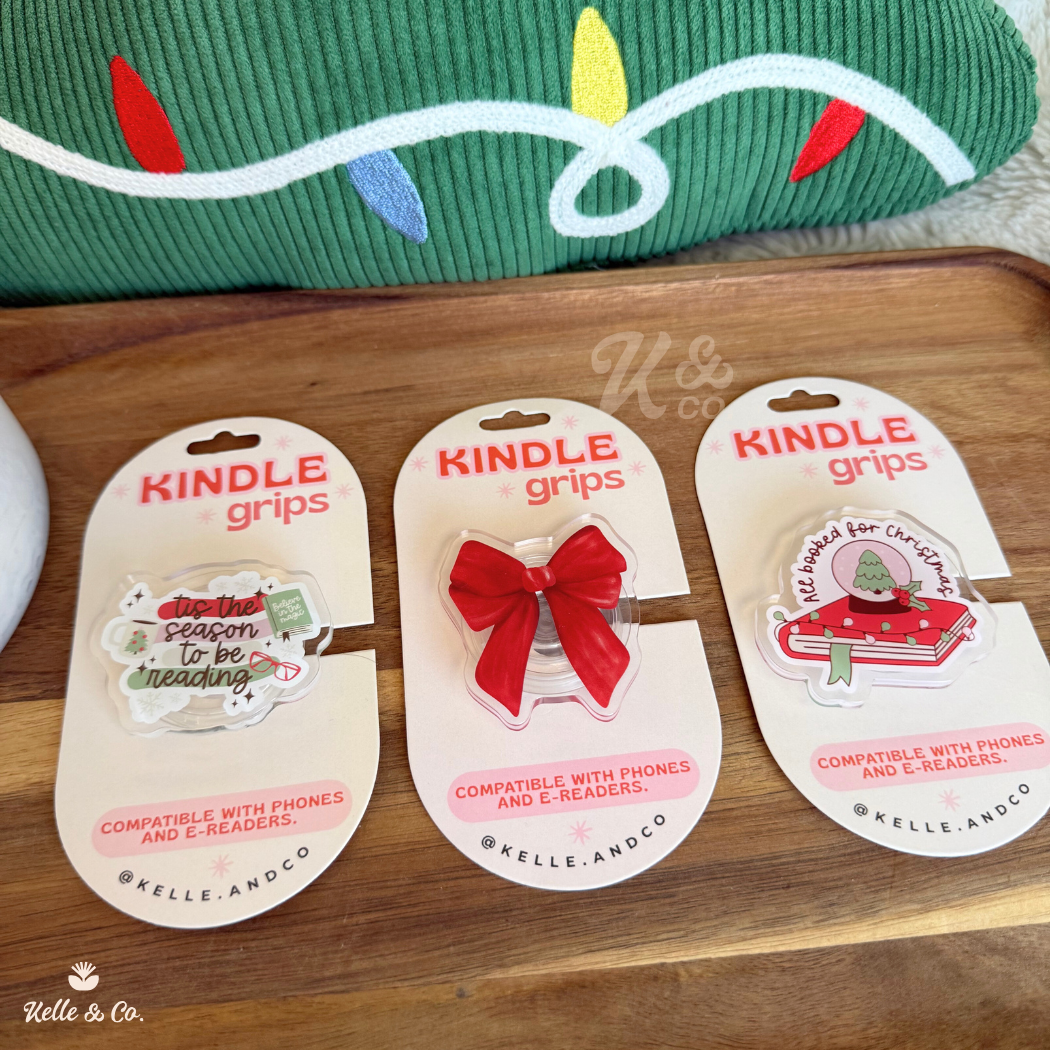 Three Christmas Kindle Grips sit on a tray, featuring a holiday book, red bow, and cat-in-a-box designs. “Christmas Kindle Grip | Christmas Book | Kindle Case”—a perfect festive gift for book lovers and trending BookTok fans.
