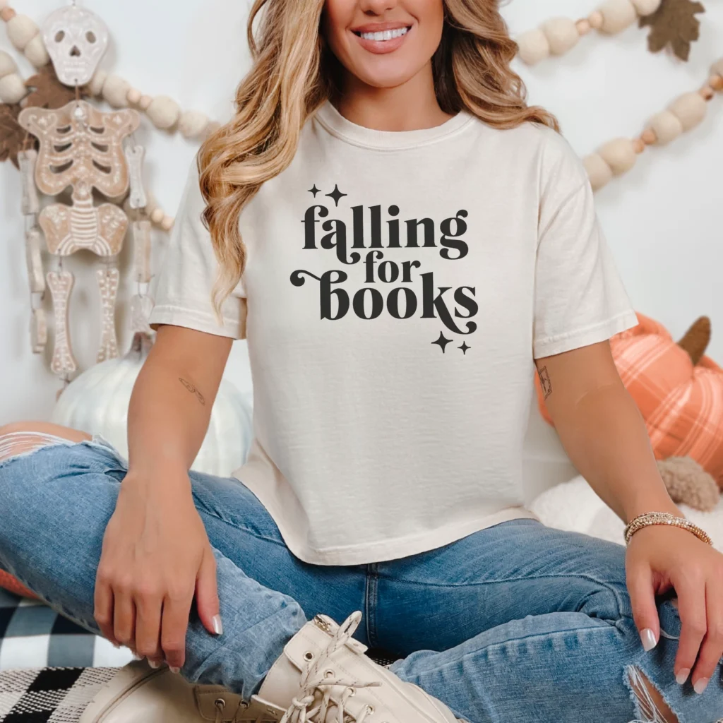 A smiling woman sits cross-legged in the Falling For Books Ivory Comfort Colors Boxy Cropped Shirt, surrounded by autumn decor and cozy pillows—an inviting spot to enjoy viral book recommendations or trending romance novels.
