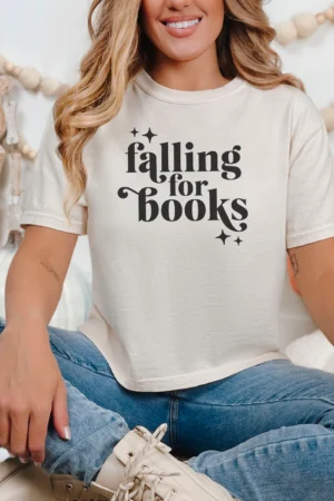 A smiling woman sits cross-legged in the Falling For Books Ivory Comfort Colors Boxy Cropped Shirt, surrounded by autumn decor and cozy pillows—an inviting spot to enjoy viral book recommendations or trending romance novels.