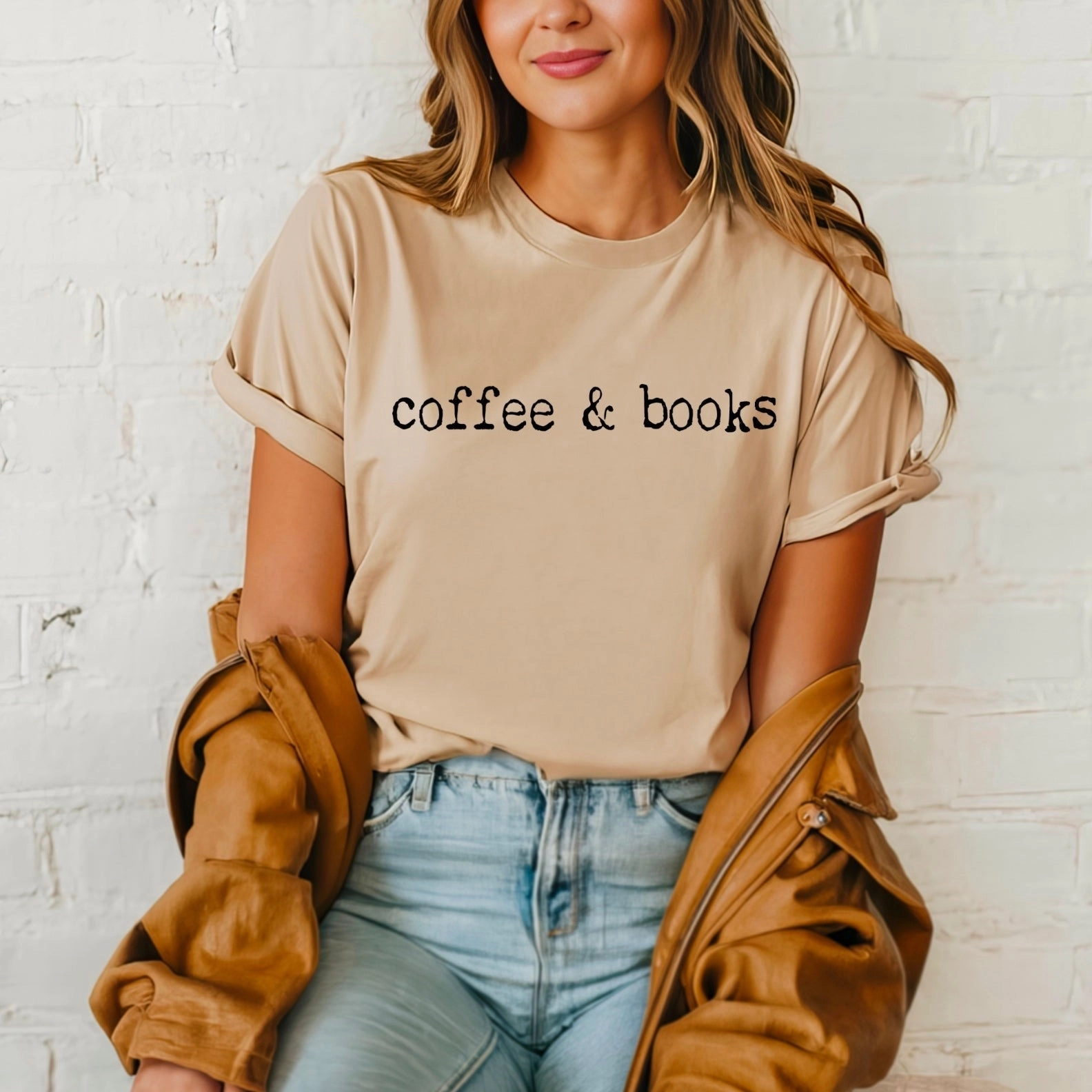 A woman with wavy hair wears the Coffee & Books Tee—beige with black text—as she sits against a white brick wall, holding a tan jacket. Her look is ideal for fans of Contemporary Fiction or Trending BookTok reads.
