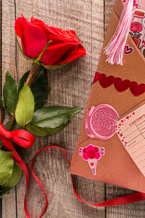 An envelope decorated with red ribbon, heart trim, and stickers sits on a wooden surface next to a single red rose and scattered red ribbon. The envelope contains bookmarks and a card labeled “Confidential Romance.”.