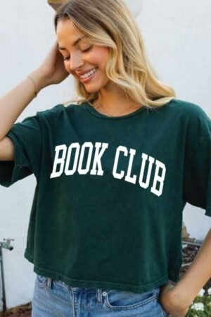 A smiling woman stands outside wearing the Book Club Cropped Mineral Graphic Top in dark green with "BOOK CLUB" text, paired with light blue jeans—ideal for fans of bestselling paperback romance novels.