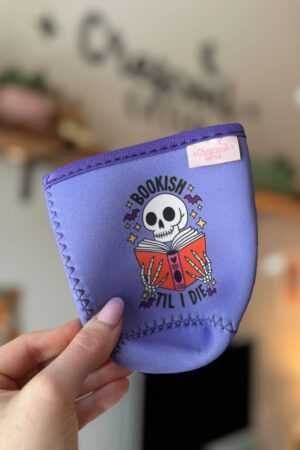 A hand holds the Bookish Till I Die Iced Coffee Sleeve, featuring a skeleton reading a red book with the phrase "Bookish Til I Die"—ideal for fans of contemporary fiction or trending book picks. Blurred shelves and plants are seen in the background.