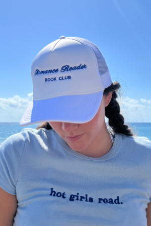 A woman in a white Romance Reader Trucker Hat and a light blue “hot girls read.” shirt stands on a sunny beach, gazing down with the ocean behind her—ideal for fans of bestselling paperbacks and viral book recs.
