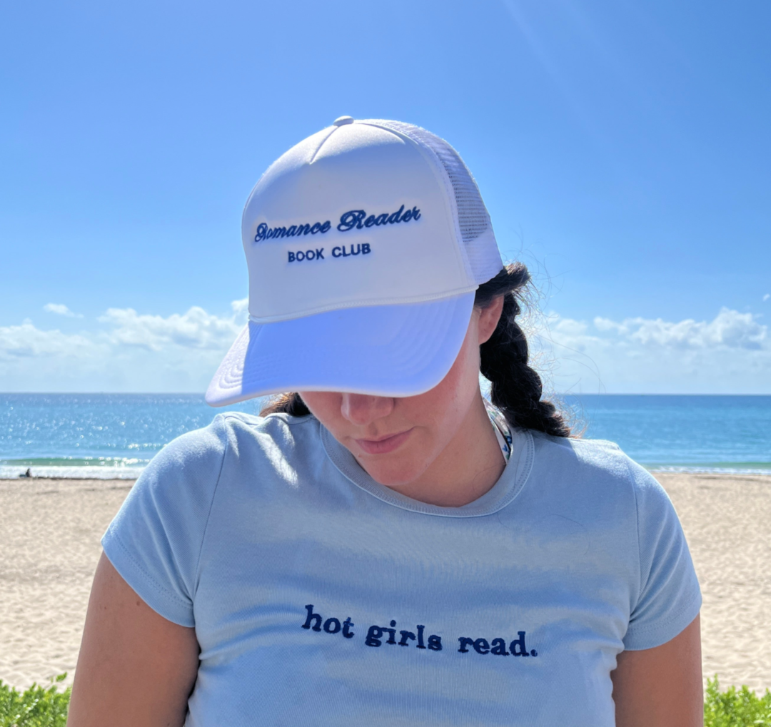 A woman in a white Romance Reader Trucker Hat and a light blue “hot girls read.” shirt stands on a sunny beach, gazing down with the ocean behind her—ideal for fans of bestselling paperbacks and viral book recs.