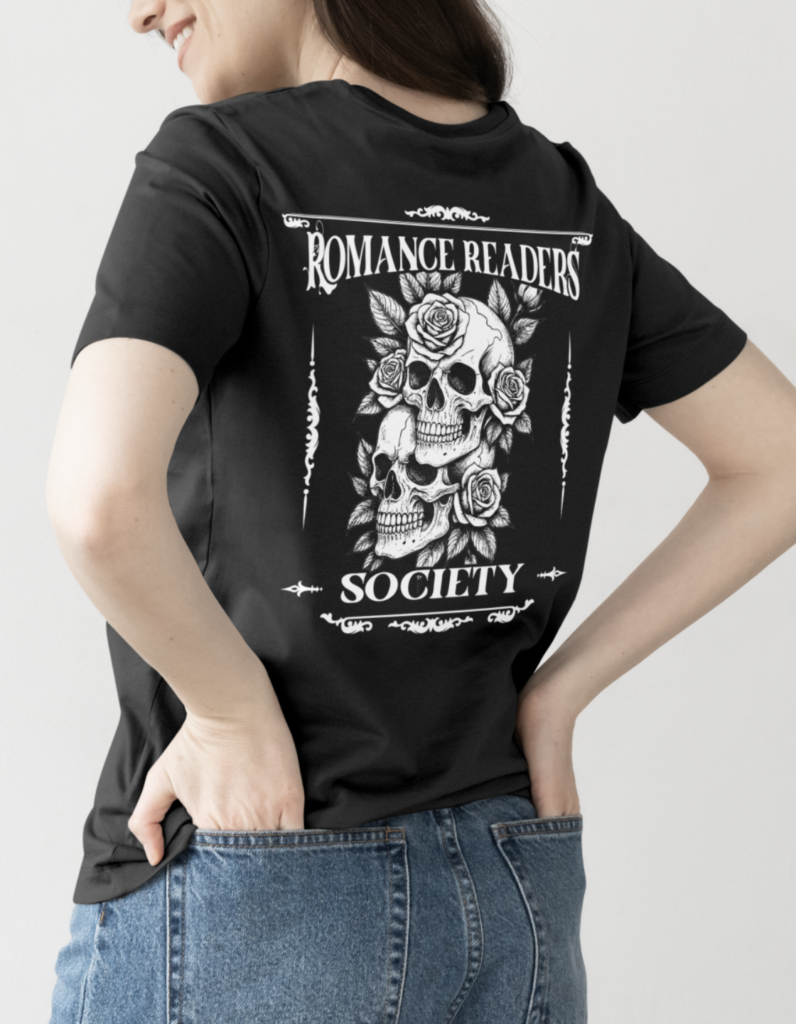 Wearing the black Romance Readers Society Tee with a white graphic of two skulls, roses, and “Romance Readers Society” text on the back, a person stands with one hand in their jean pocket.