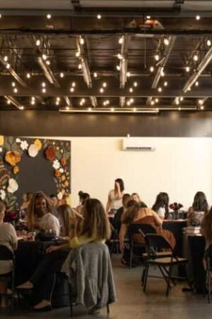 Flourish Books book club gathers around tables in a modern, well lit room adorned with string lights and a colorful mural, visible through an open doorway at the February Blush Club.