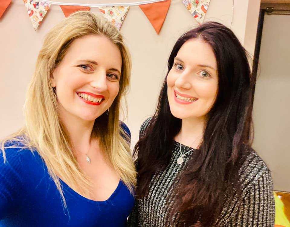 Authors Peckham and Valenti smile indoors with festive bunting behind them. One wears a blue top, the other stripes—perhaps celebrating their love for Zodiac Academy 2: Ruthless Fae by Caroline Peckham & Susanne Valenti (Paperback).