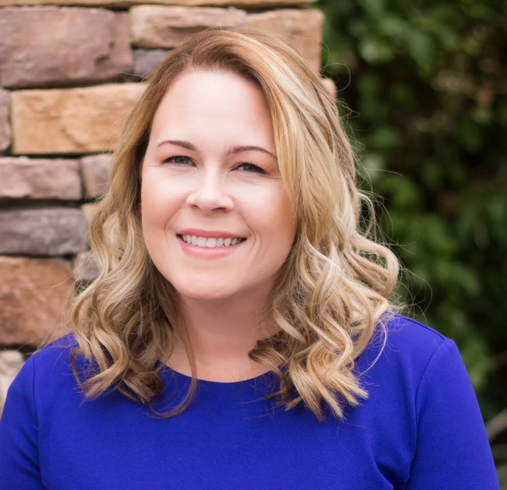 Author Rebecca Jenshak in a royal blue top smiles before a stone wall and greenery, echoing the heroine from "Lucky Shot" (Moonshot Hockey) by Rebecca Jenshak (Paperback).