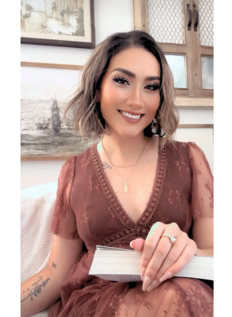 KM Moronova with wavy brown hair in a mauve lace V neck dress, gold necklace, and ring holds "Your Knife, My Heart (Standard Edition) (Dark Forces) by K M Moronova." She has visible arm tattoos and sits before framed wall art.