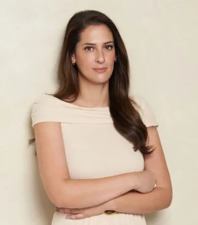 Elle Kennedy with long brown hair in a light beige dress stands against a plain background, arms crossed and neutral faced—capturing the understated elegance of "Love Song (Deluxe Edition)" by Elle (Paperback).