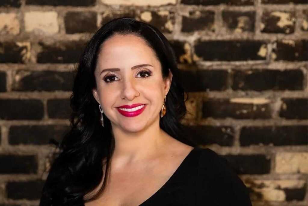 Author Abby Jimenez with long dark hair and bright pink lipstick smiles at the camera in front of a rustic brick wall, wearing a black top and gold earrings, embodying the spirit of The Night We Met (Deluxe Edition) by Abby Jimenez.