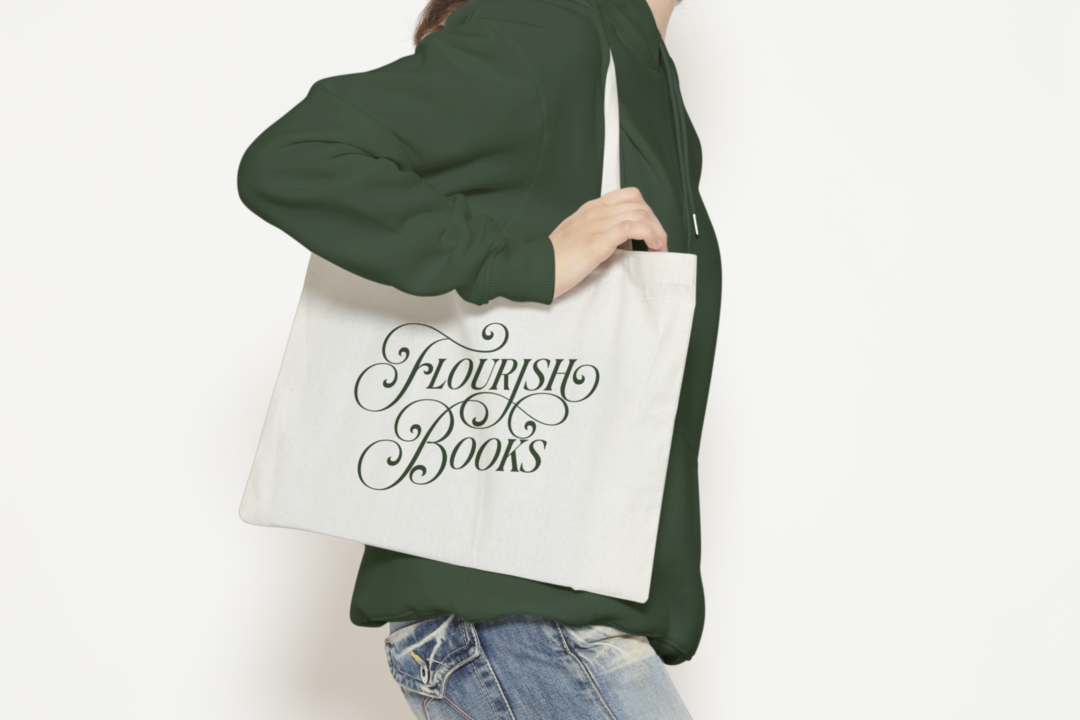 A person in a green hoodie and jeans holds the Flourish Tote, featuring ornate green script reading "Flourish Books," against a plain white background.