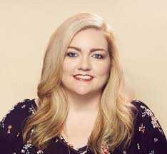 Author Colleen Hoover with long, wavy blonde hair wears a floral top against a beige background, inspired by "Verity" by Colleen Hoover.