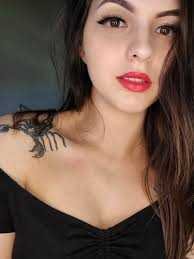 Author HD Carlton with long dark hair and red lipstick wears a black off the shoulder top. Inspired by "Does It Hurt? by H. D. Carlton," she reveals a bold dragon tattoo sprawling across her left shoulder.