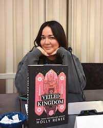 Author Holly Renee with long dark hair and a gray sweatshirt smiles with her chin resting on her hands, sitting behind a signed Special Edition of Cowboy Casual by Holly Renee. Light vertical blinds are in the background.