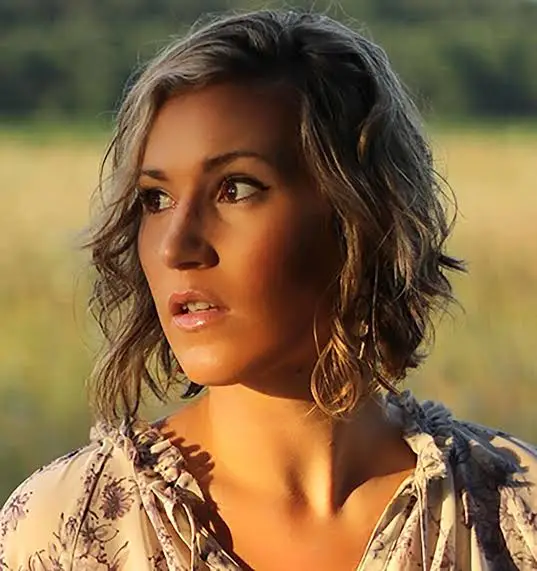 Author Navessa Allen with wavy, shoulder length blonde hair gazes to the side in soft sunlight, wearing a floral top and channeling the effortless style found in Game On by Navessa Allen (Into Darkness, 3 of 3), against a blurred natural backdrop.