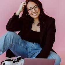 Author Kat Singleton with long dark hair and glasses sits cross legged before a pink backdrop, smiling as she reads More Than Friends – by Kat Singleton (Paperback), with a pink laptop and white coffee mug nearby.