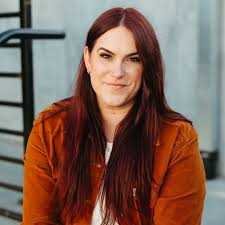 Liz Tomforde with long, straight reddish brown hair and fair skin smiles slightly while reading "Mile High (Windy City) by Liz Tomforde (Paperback)" outdoors—a perfect Mile High vibe.