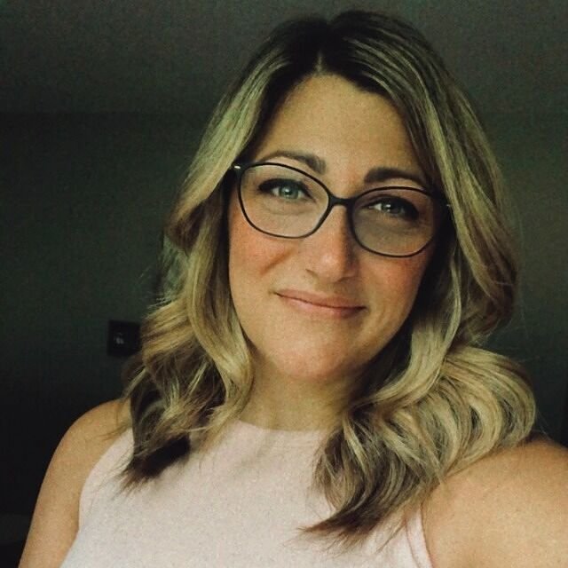 A woman with wavy, shoulder length blonde hair and glasses smiles softly at the camera, wearing a sleeveless light pink top—a cozy scene inspired by "Bourbon & Proof (Deluxe Edition)" by Victoria Wilder (Paperback).