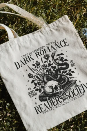 The Dark Romance Gothic Skull Tote sits on grass, featuring "Proud member of the Dark Romance Readers Society. Enemies to lovers. Est. 2021. Trigger warnings welcome," with floral, snake, and skull artwork; a candle and ornaments nearby.
