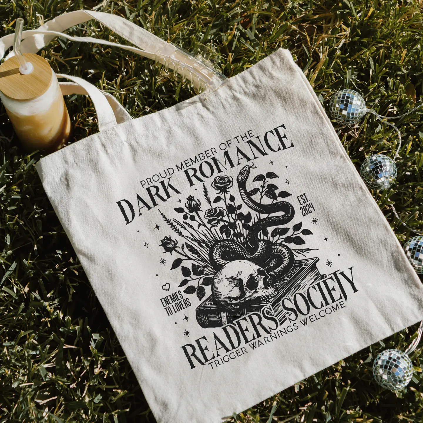 The Dark Romance Gothic Skull Tote sits on grass, featuring "Proud member of the Dark Romance Readers Society. Enemies to lovers. Est. 2021. Trigger warnings welcome," with floral, snake, and skull artwork; a candle and ornaments nearby.