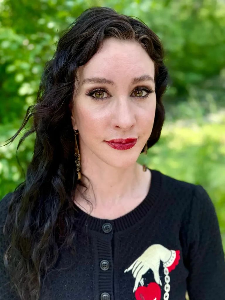 Author Brynne Weaver with long wavy dark hair and green eyes stands outdoors among green foliage, wearing a black sweater inspired by "Harvest Season" by Brynne Weaver, with a hand and heart motif, red lipstick, and dangling earrings.