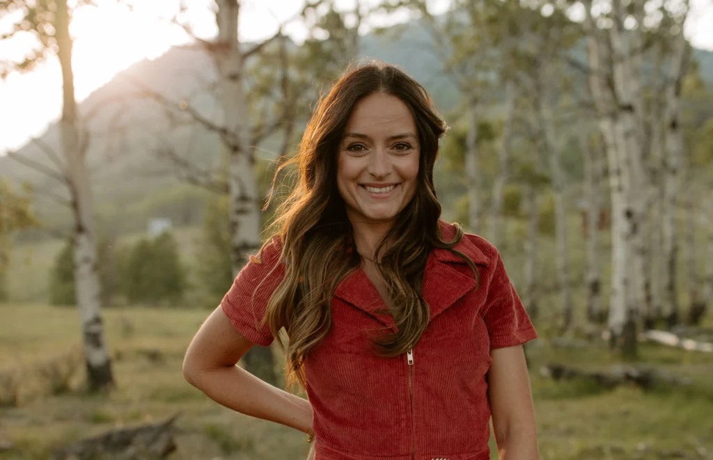 Author Elsie Silver with long brown hair smiles outdoors in a short-sleeved red dress, basking in sunlight—evoking the May Blush Club: Cowboy Romance & "Flawless" at Beach Vodka vibe under open skies.