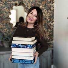 Kerri Maniscalco with long brown hair and a dark sweater stands indoors holding a tall stack of hardcover books, including Kingdom of the Wicked by Kerri Maniscalco. A floral patterned mirror and light wall are in the background.